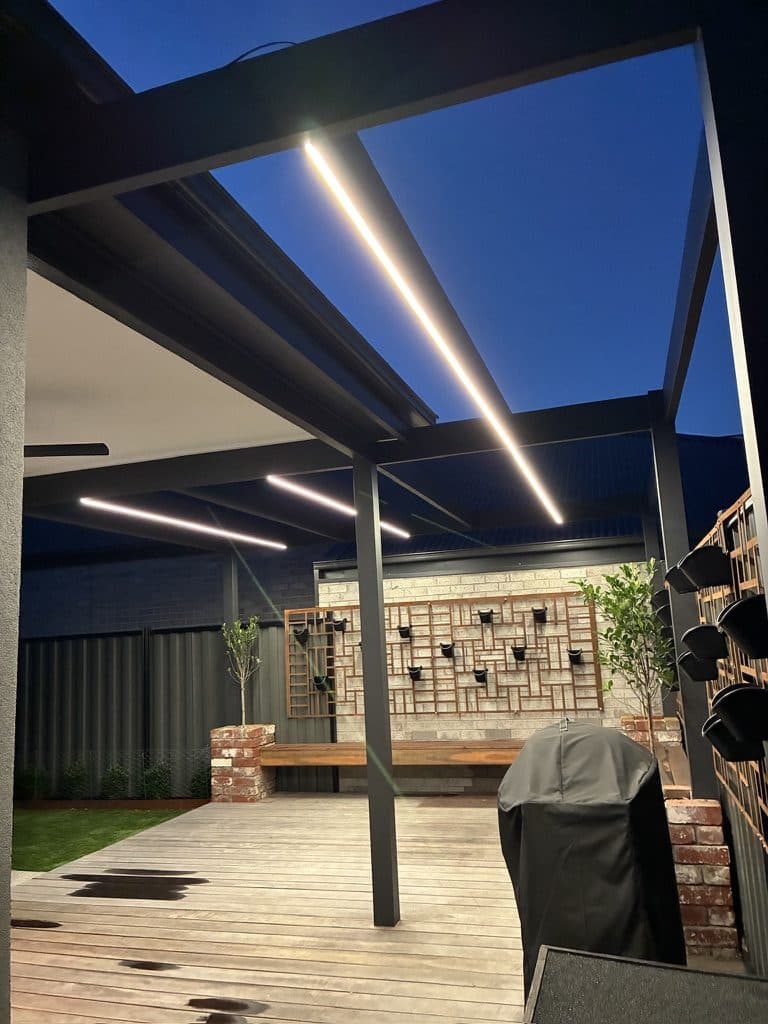 A covered alfresco at dusk, lit by strip lights in the roof, with timber screens and bench seating around a deck