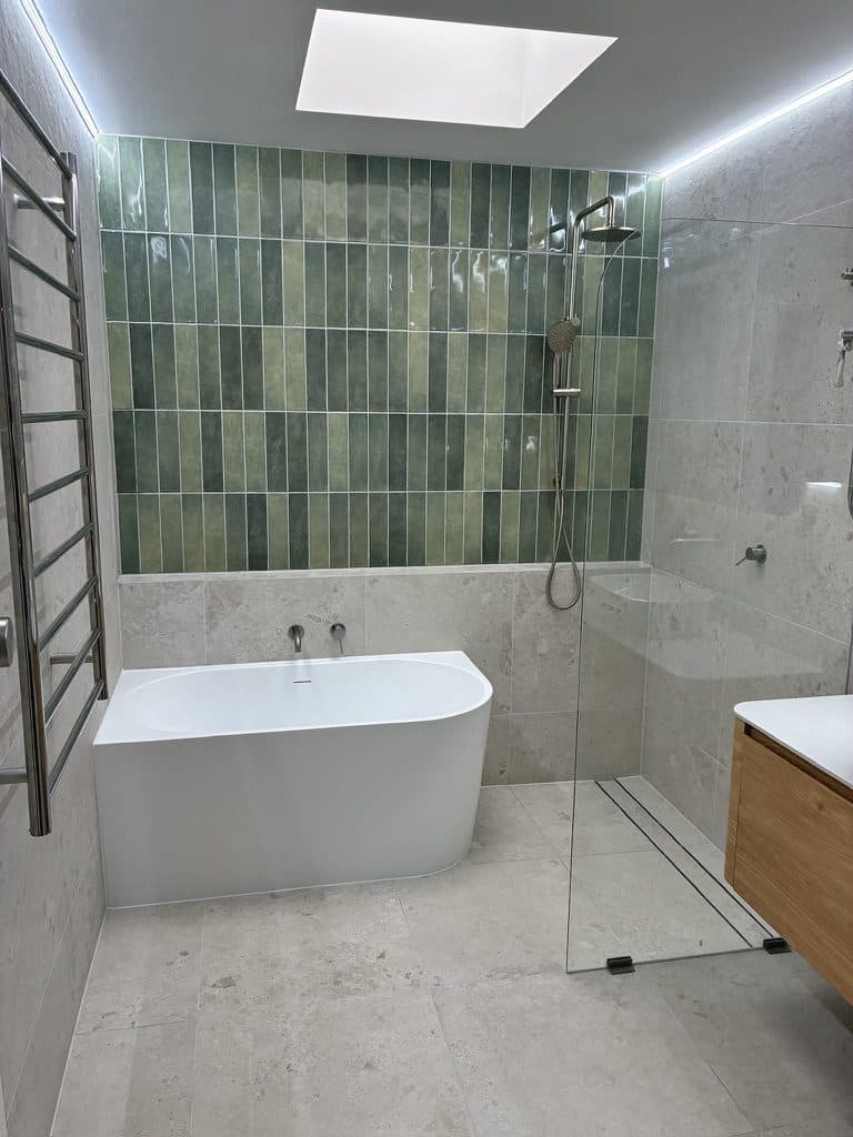 A finished bathroom with a green vertical tile wall, a freestanding bath, a frameless shower screen and a skylight