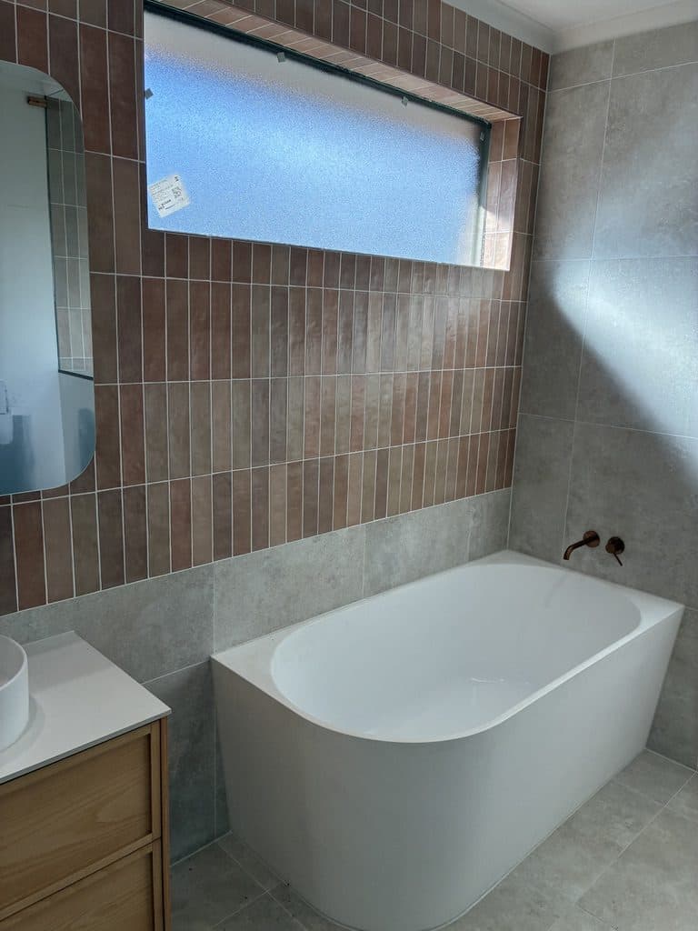 A finished bathroom with a freestanding corner bath, vertical terracotta tiles and a high window