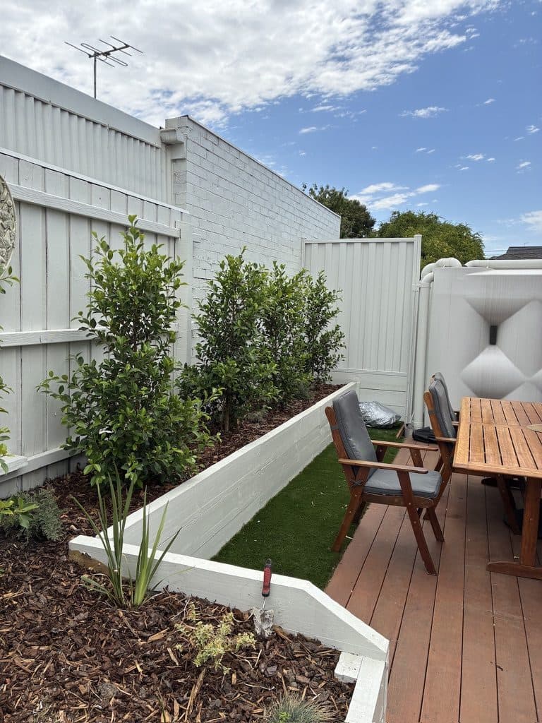 A deck beside a raised white planter with shrubs and synthetic lawn