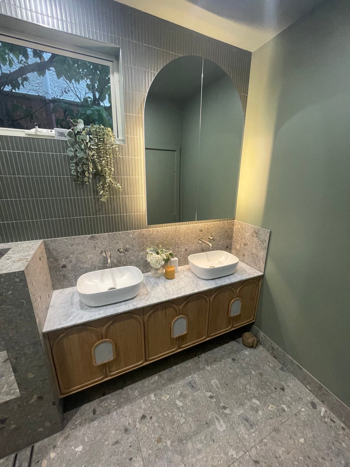 A finished bathroom with terrazzo tiles, a green wall, an arched mirror recess and a timber double vanity