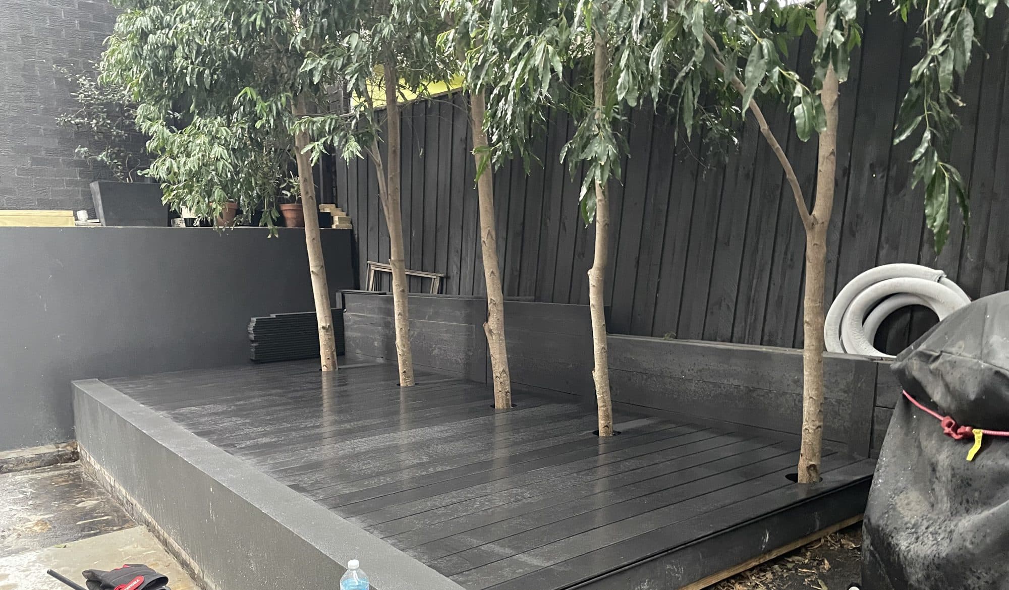 A new dark composite deck built inside a raised rendered courtyard bed, trees along the fence