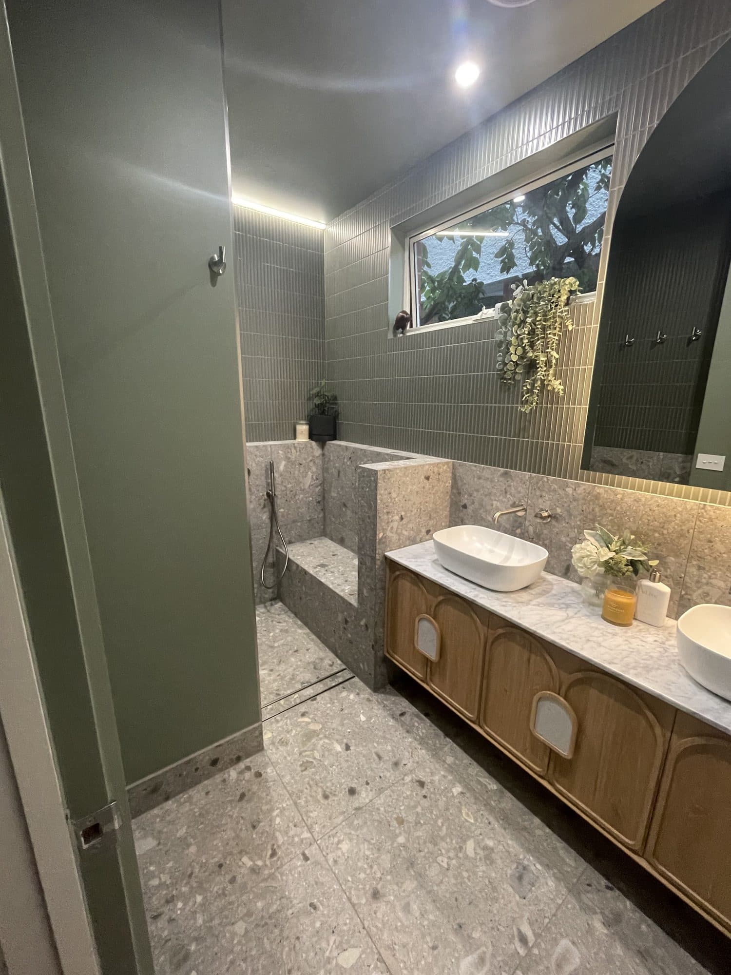 A finished bathroom with a timber vanity under a marble top, terrazzo floor and green tiled shower