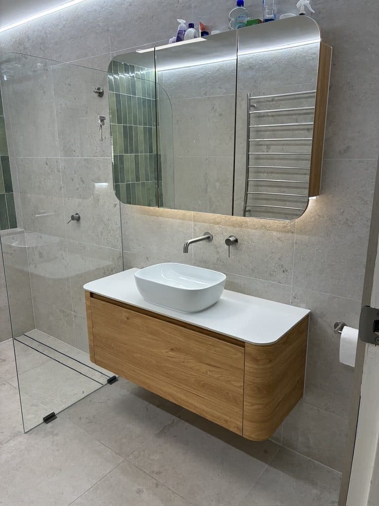 A curved timber vanity with a basin and a lit mirror cabinet