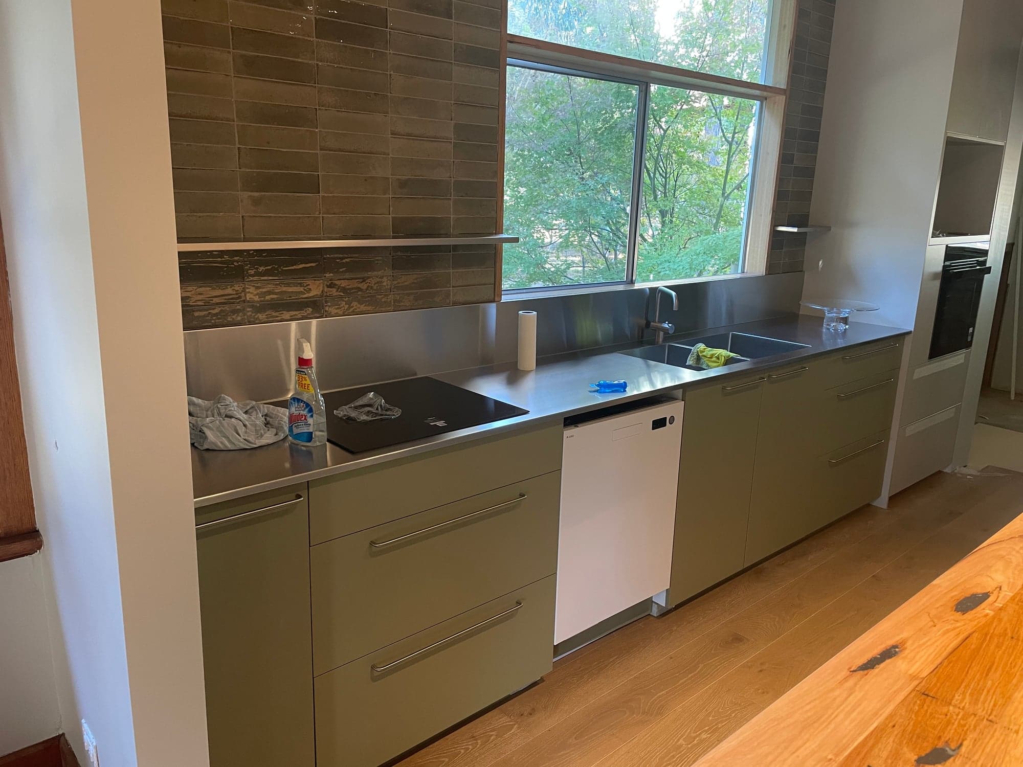 The kitchen along the window wall: sage-green cabinets, a stainless benchtop and a tiled splashback